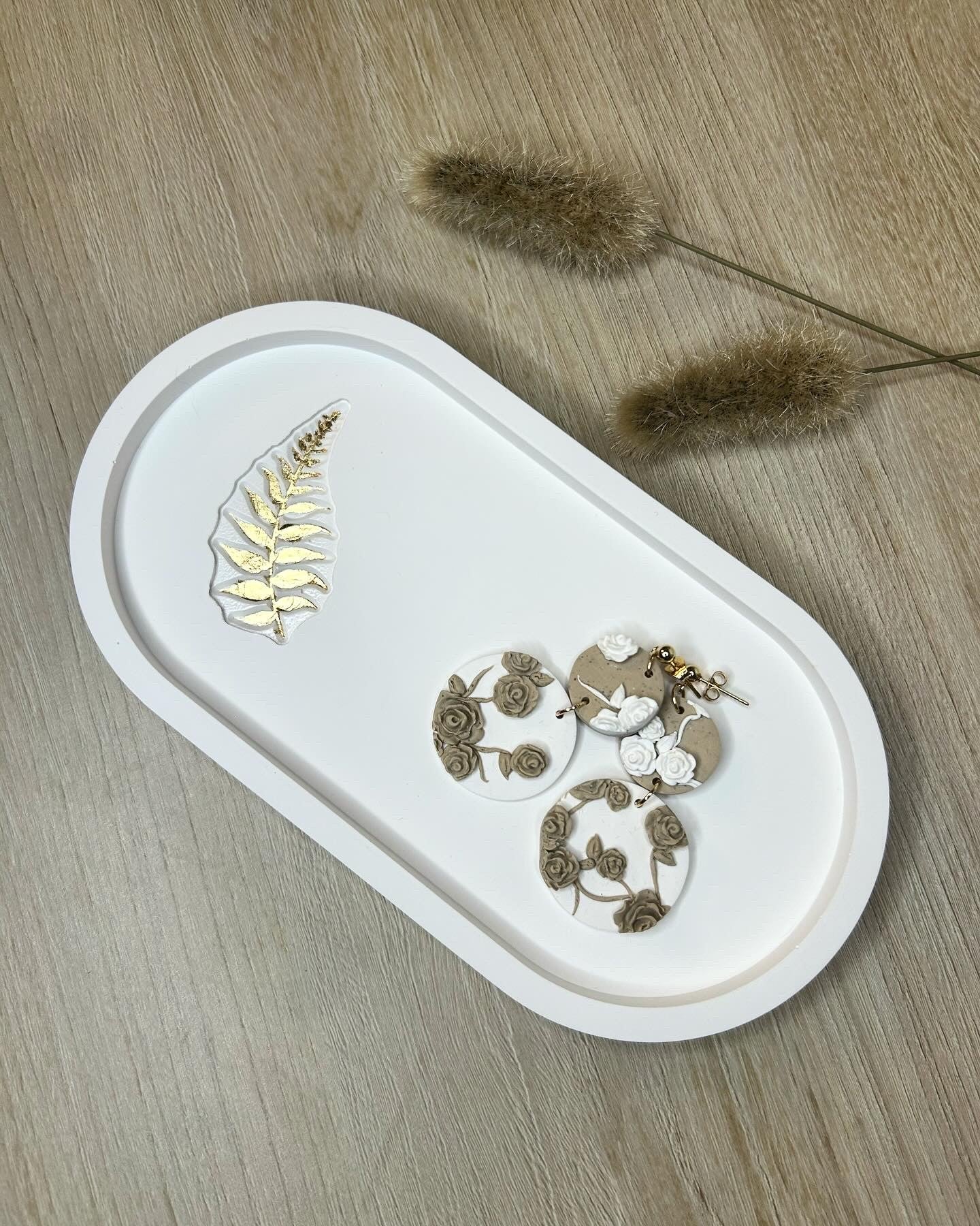 Handmade Home Accessories - A white oval trinket tray with a gold leaf fern leaf detail. It has a pair of cream and beige floral earrings on and is sat on a background of light oak wood with some pampas grass accents.